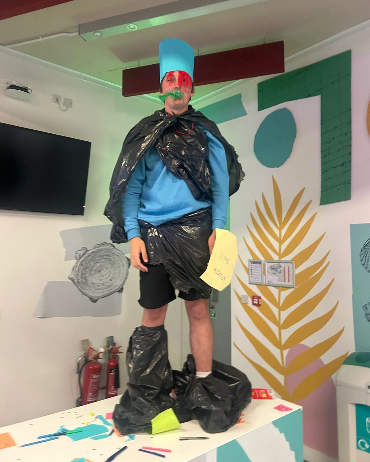 Alexander Burns in a costume of black bin bags and a tall blue paper hat, with face paint, at a bin bag fashion show