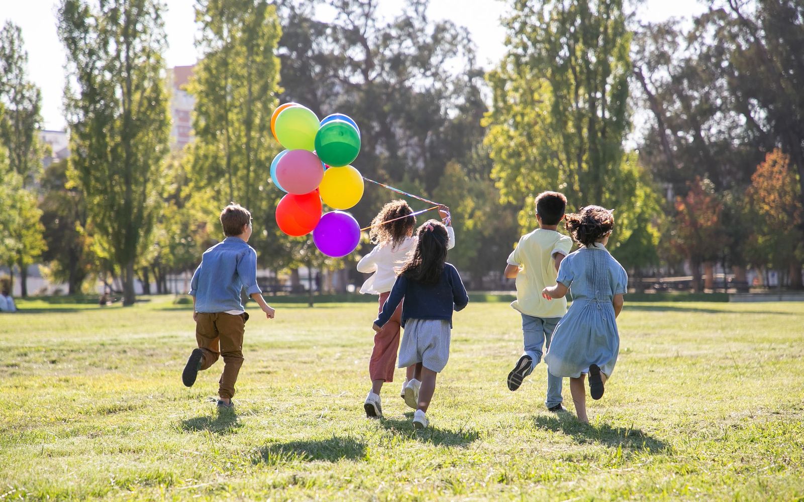 Children running across a park with a bunch of balloons, seen from behind